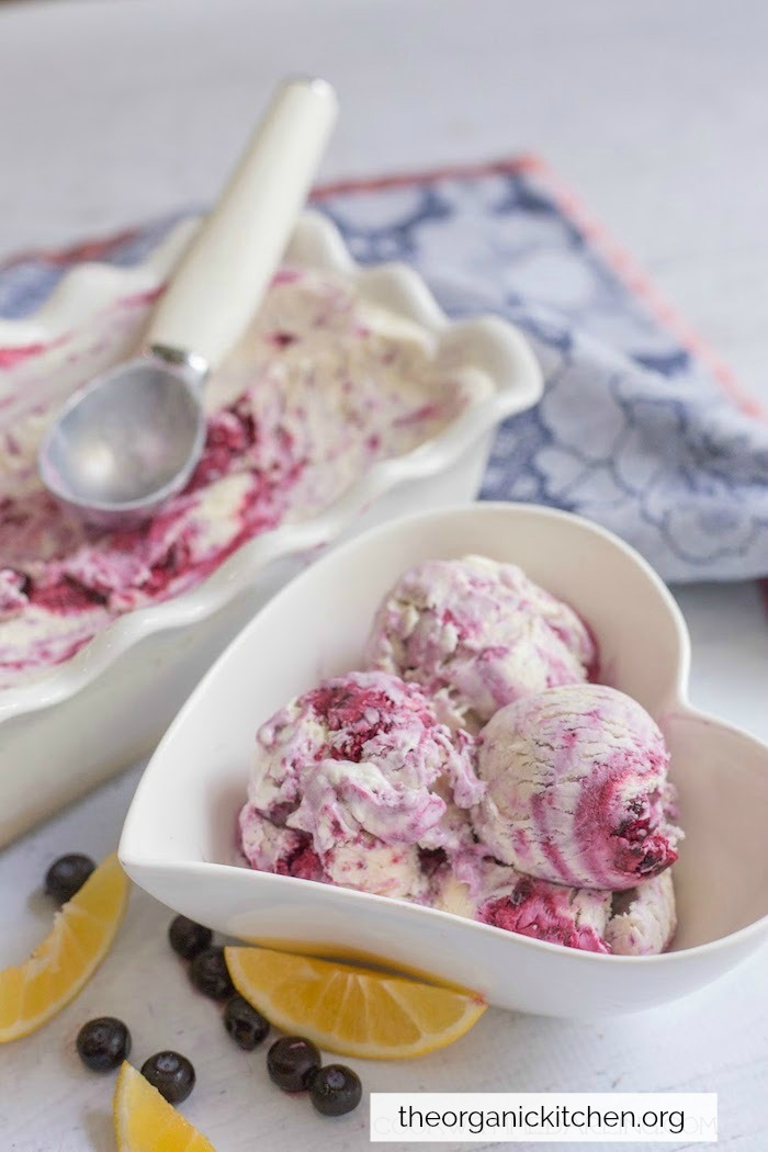 A bowl and a dish of Blueberry Swirl No Churn Ice Cream with an ice cream scoop and a dish towel in background. Five No Churn Ice Cream Recipes