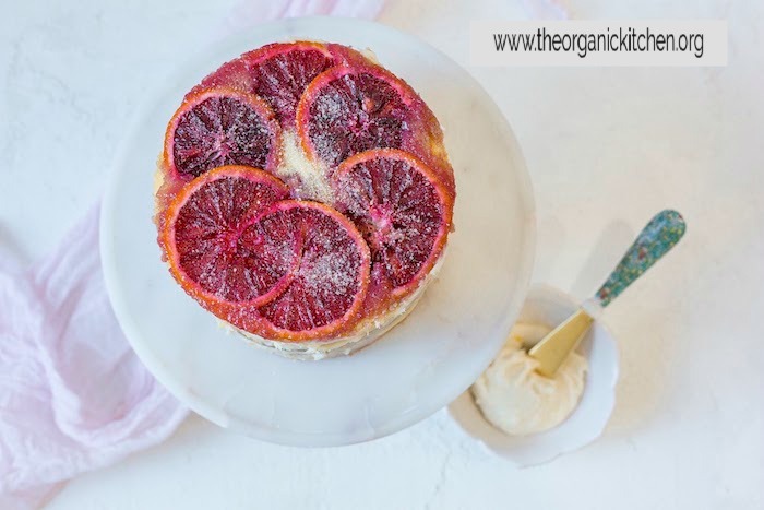 A small layer cake topped with blood oranges on white marble cake stand