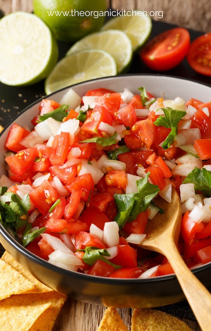 Fresh Pico de Gallo in bowl with wooden spoon with limes and tomatoes in background