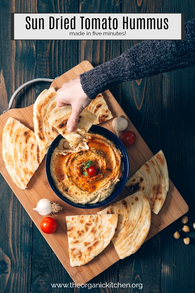 A woman's hand dipping pita bread into Sun Dried Tomato Hummus