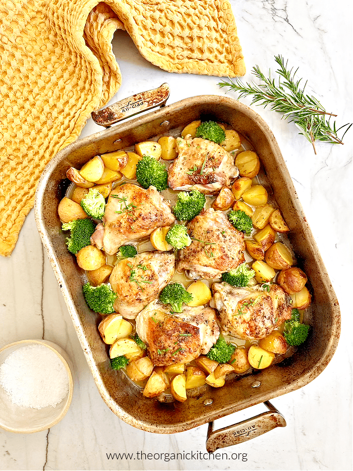 Lemon Chicken with Roasted Potatoes and broccoli in roasted pan on white counter surrounded by yellow dish towel and rosemary sprigs