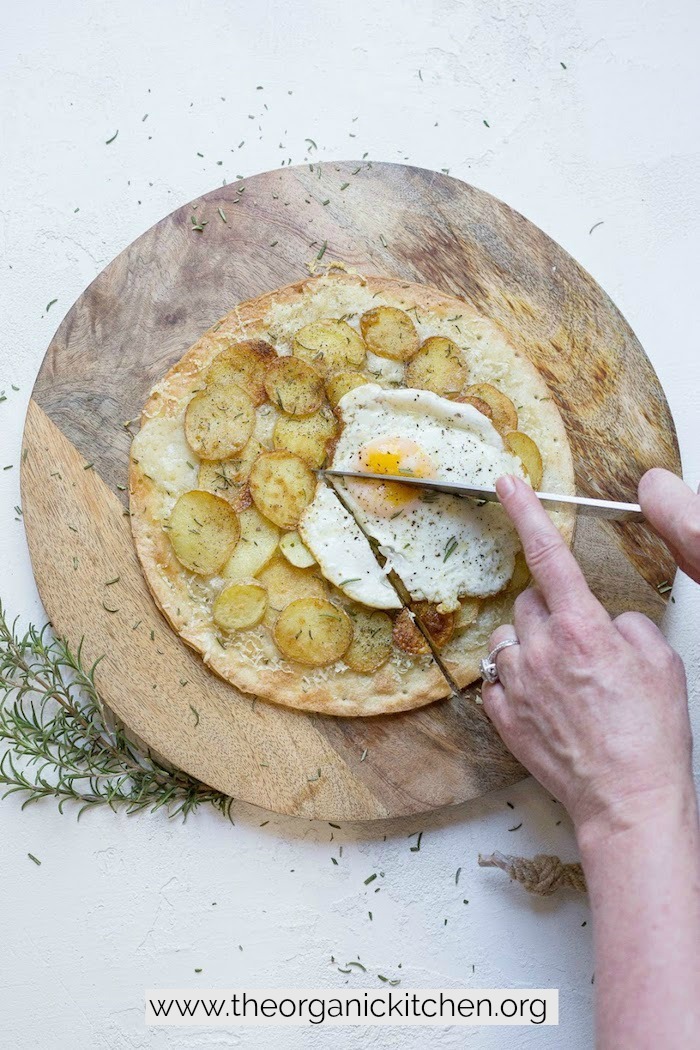 Hands holding a knife and cutting a slice of Rosemary Potato Flatbread with Cauliflower Crust Option 