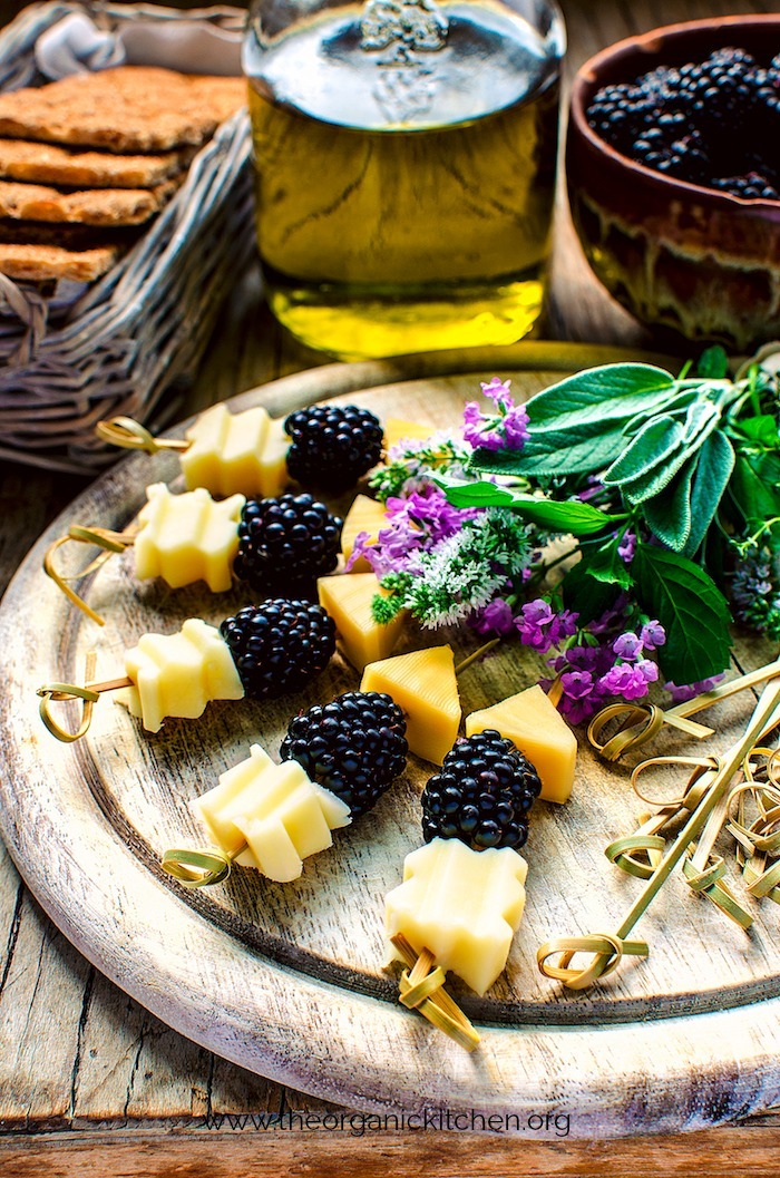 Blackberry and Aged Cheese Skewers on a serving board with baskets and bowls in the background filled with crackers and fruit