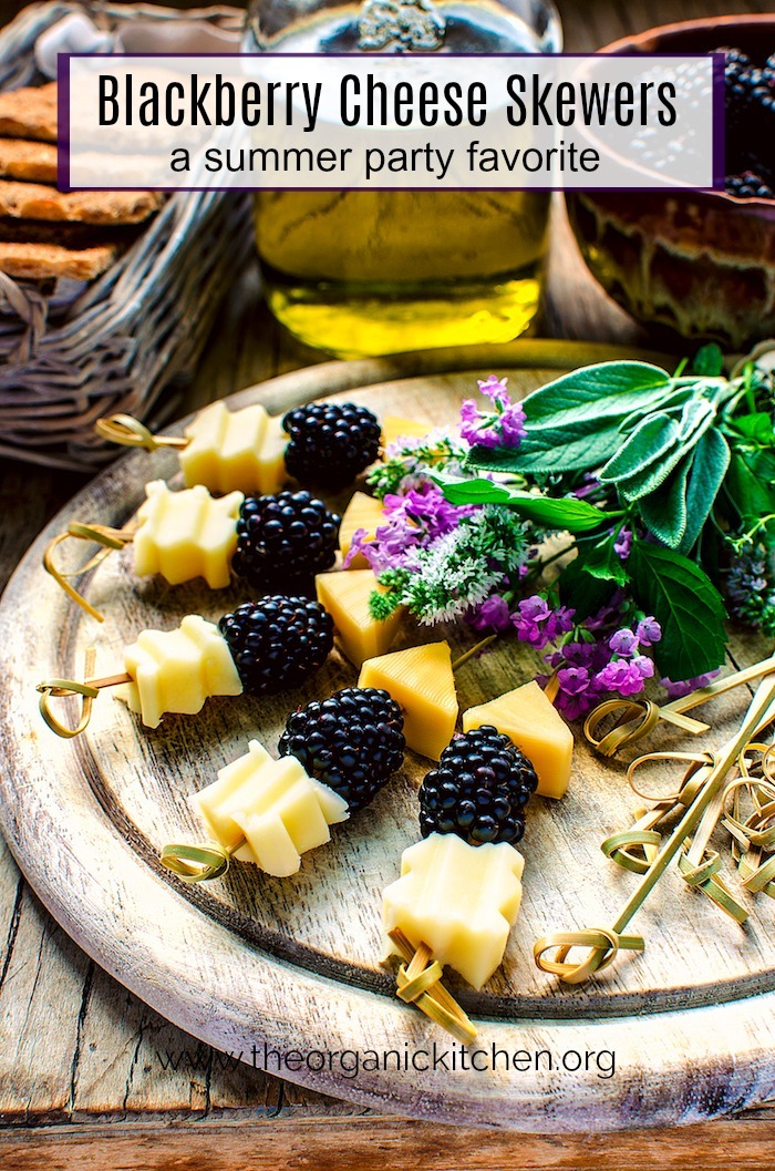 Blackberry and Aged Cheese Skewers on a round wood platter garnished with herbs and flowers