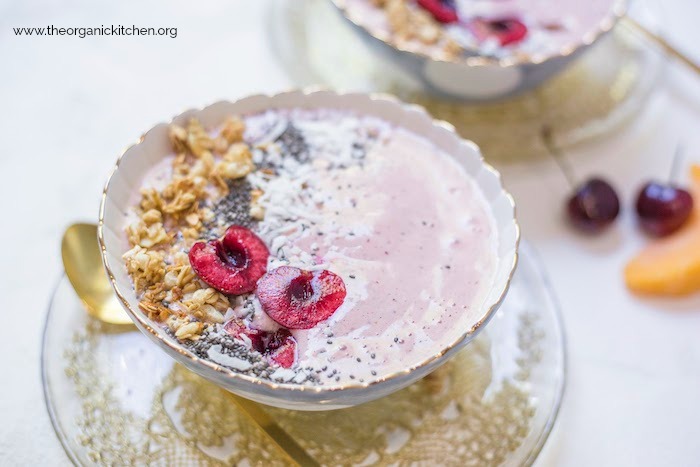 A close up of Peach Cherry Smoothie Bowls on gold laced plates topped with cherries, granola and chia