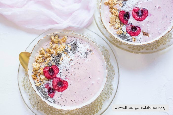 Two Peach Cherry Smoothie Bowls on gold laced plates topped with cherries, granola and chia on a white surface with pink fabric