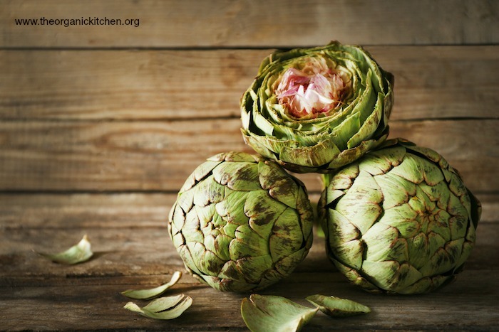 Three artichokes, one trimmed, set on a wooden table