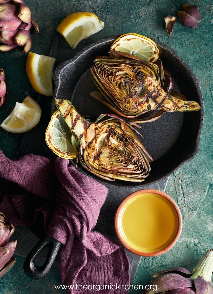 Grilled artichoke halves in a black cast iron pan with a small bowl of melted butter