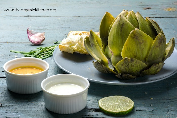 A cooked artichoke on a gray plate with small bowls of melted butter and mayonnaise 