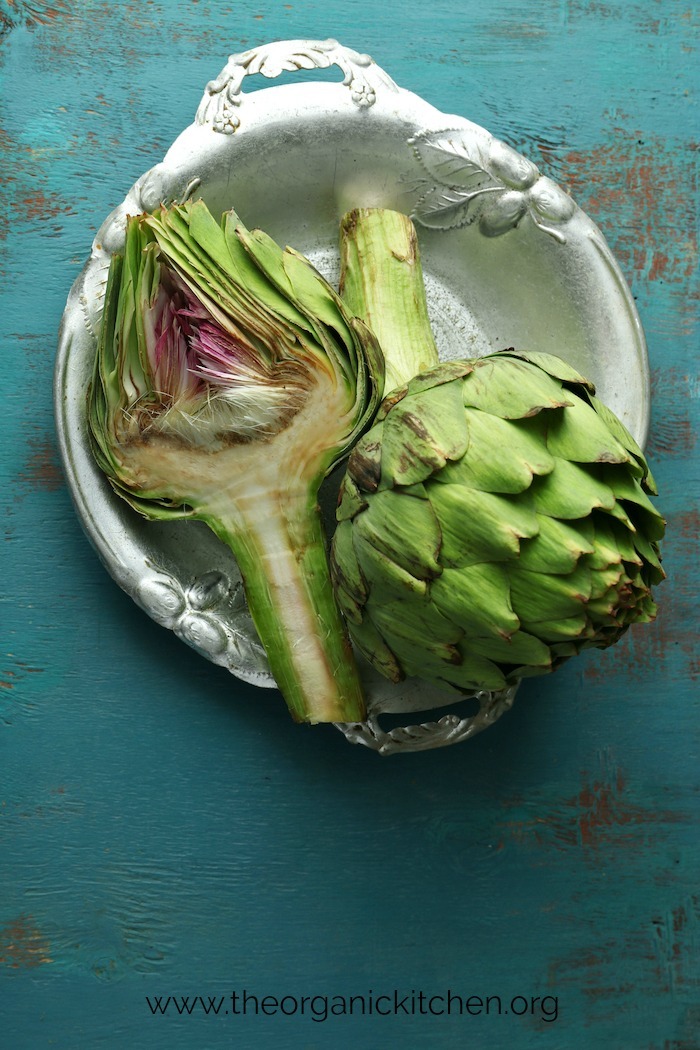 Two artichokes in a white bowl set on a turquoise table