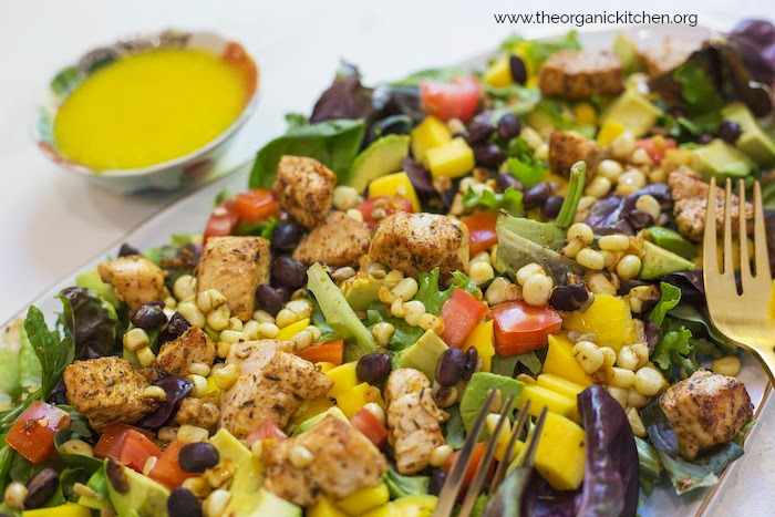 A close up of Southwest Chicken Salad with Mango Vinaigrette showing greens, corn, black beans, diced mango and a small bowl of bright yellow salad dressing