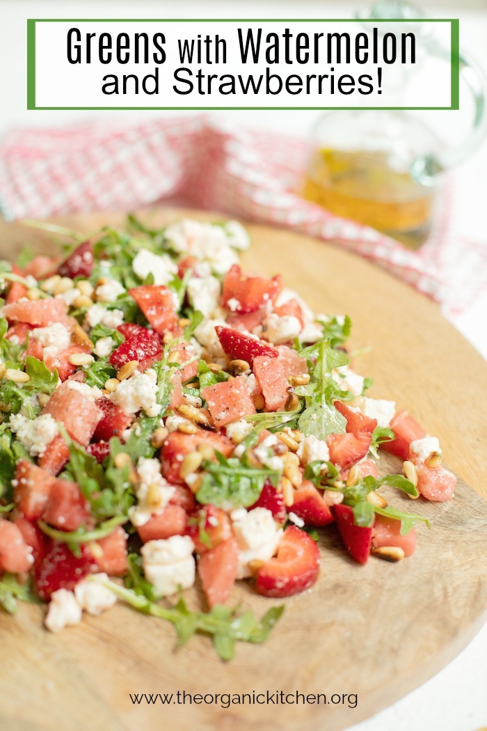 A wooden platter filled with greens with watermelon and strawberries with a bottle of salad dressing and dish towel in background