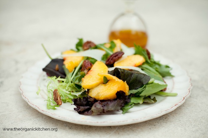 Green Salad with Fresh Peaches on a white dish set on a stone surface with a bottle of salad dressing