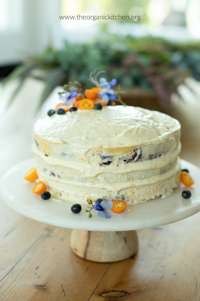 Orange Blueberry Cake with Orange Buttercream Frosting decorated with fruit and flowers on a white cake plate set on a table