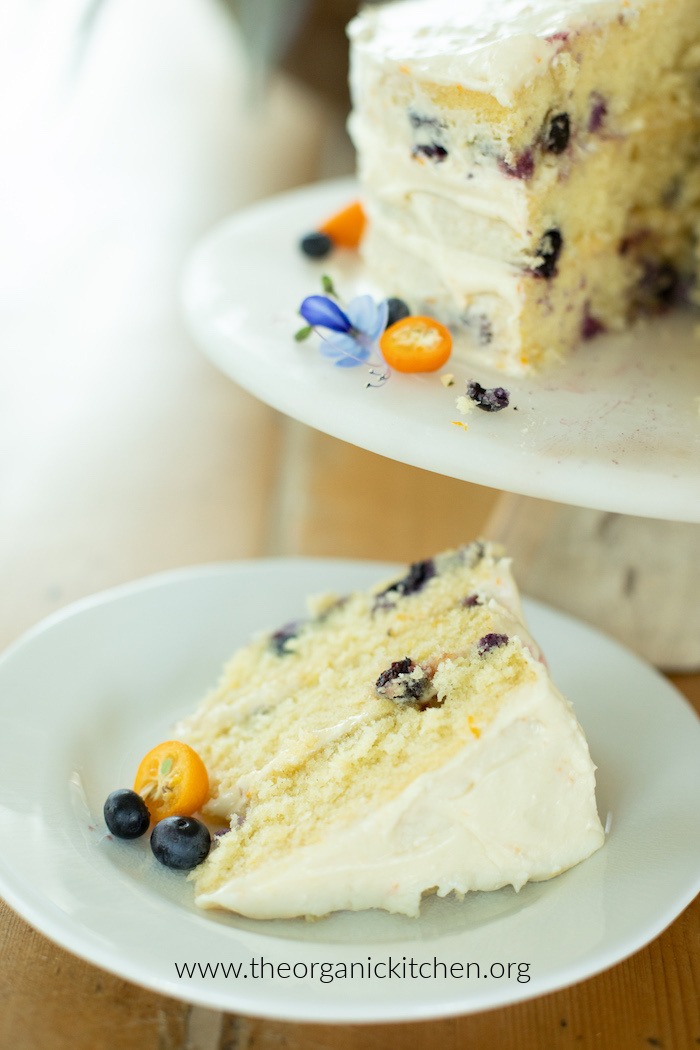 A slice of Orange Blueberry Cake on a white plate set next to a cake plate with the rest of the cake