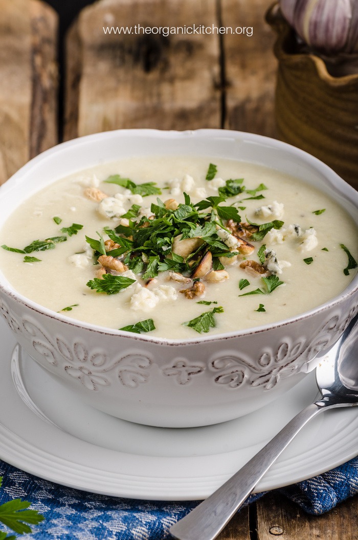 A white bowl sitting on a saucer filled with Roasted Cauliflower Soup garnished with herbs