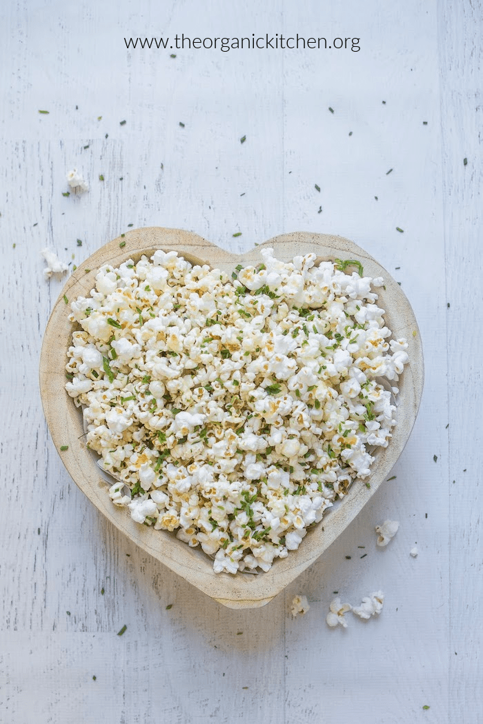 Parmesan Chive Popcorn in heart shaped bowl on white surface scattered with popcorn and chives