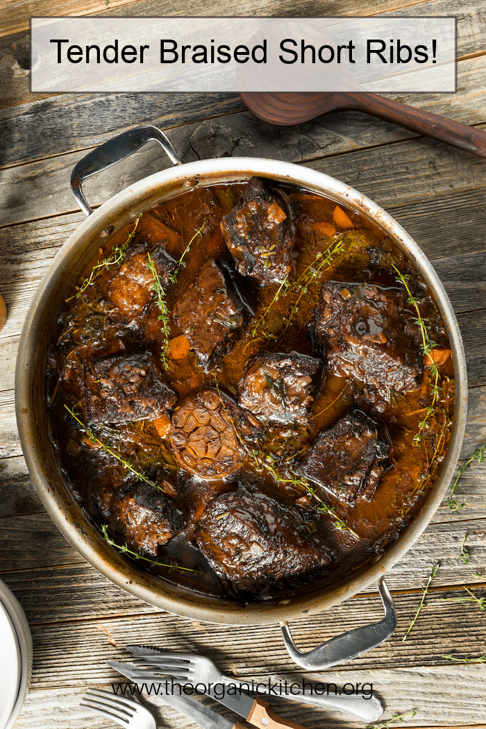 Tender Braised Short Ribs with a red wine sauce in a pot set on a table, surrounded by silverware and a wooden spoon