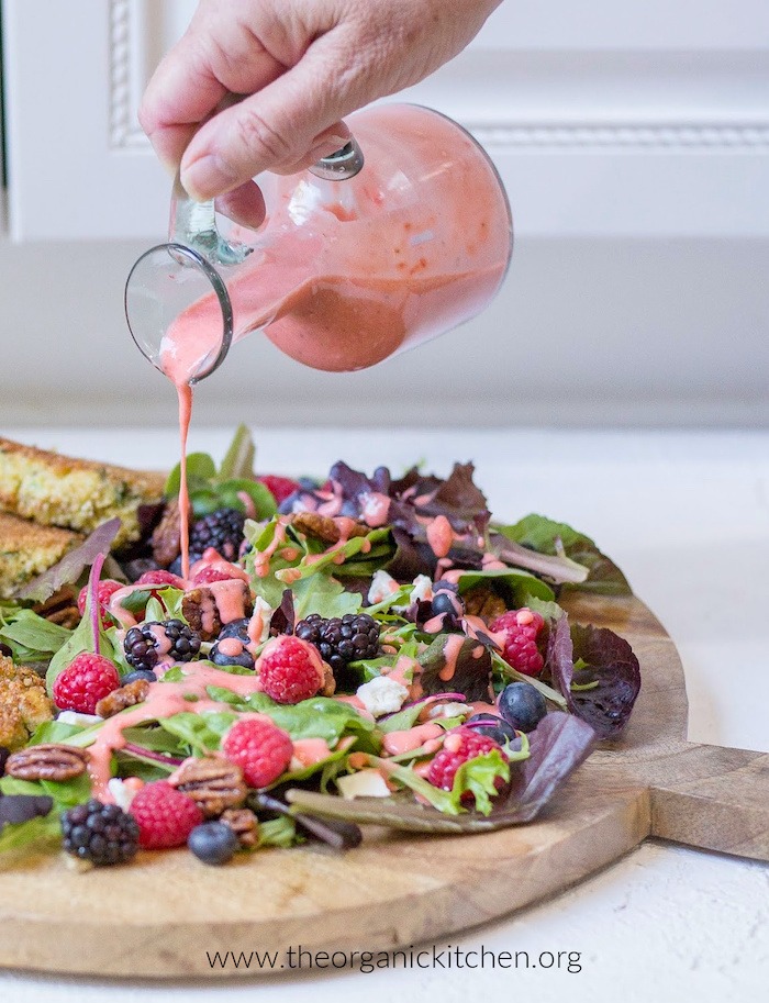 A females hand holding drizzling Creamy Strawberry Vinaigrette - Whole30/Paleo onto a fresh berry salad