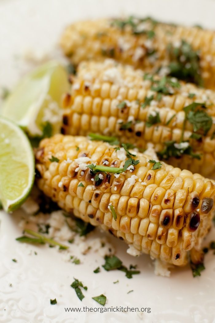 Three ears of Mexican Street Corn garnished with cilantro, lime wedges  and cheese 