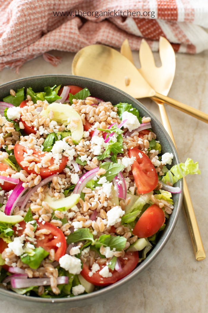 Chopped Greek salad in a grey bowl with gold serving utensils