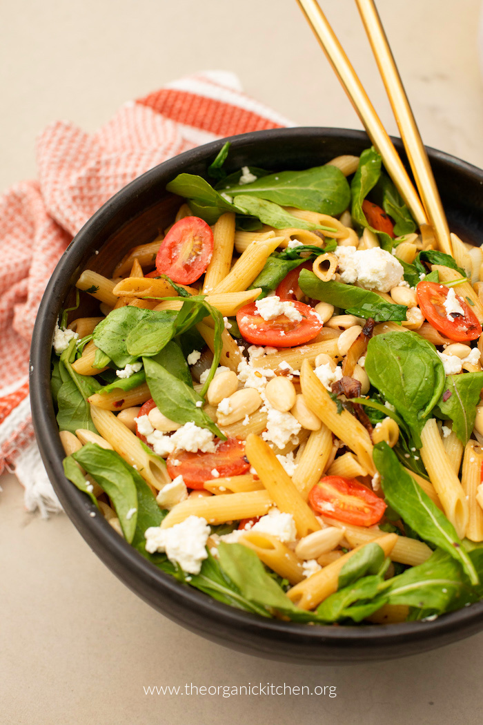 Penne Pasta with Tomatoes and Baby Arugula in black bowl with red and white dish towel in background