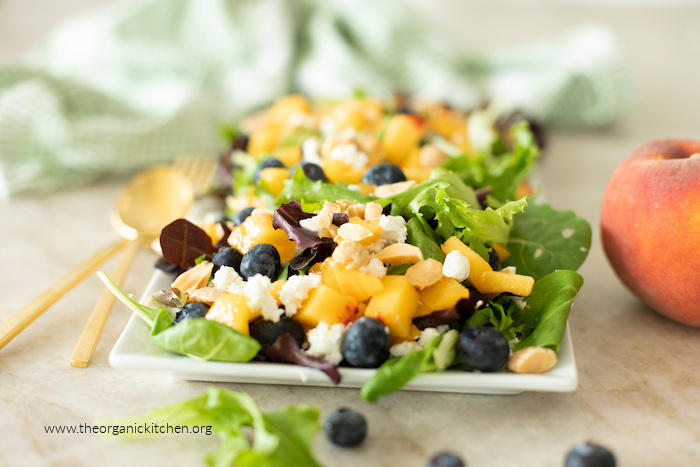 A peach salad with blueberries and feta on a white plate with gold serving utensils on the side