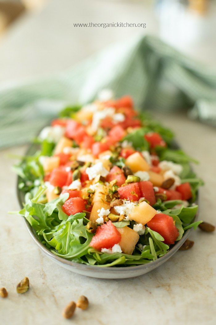 Summer Melon Salad garnished with feta and pistachios on gray platter with green and white dish towel in background