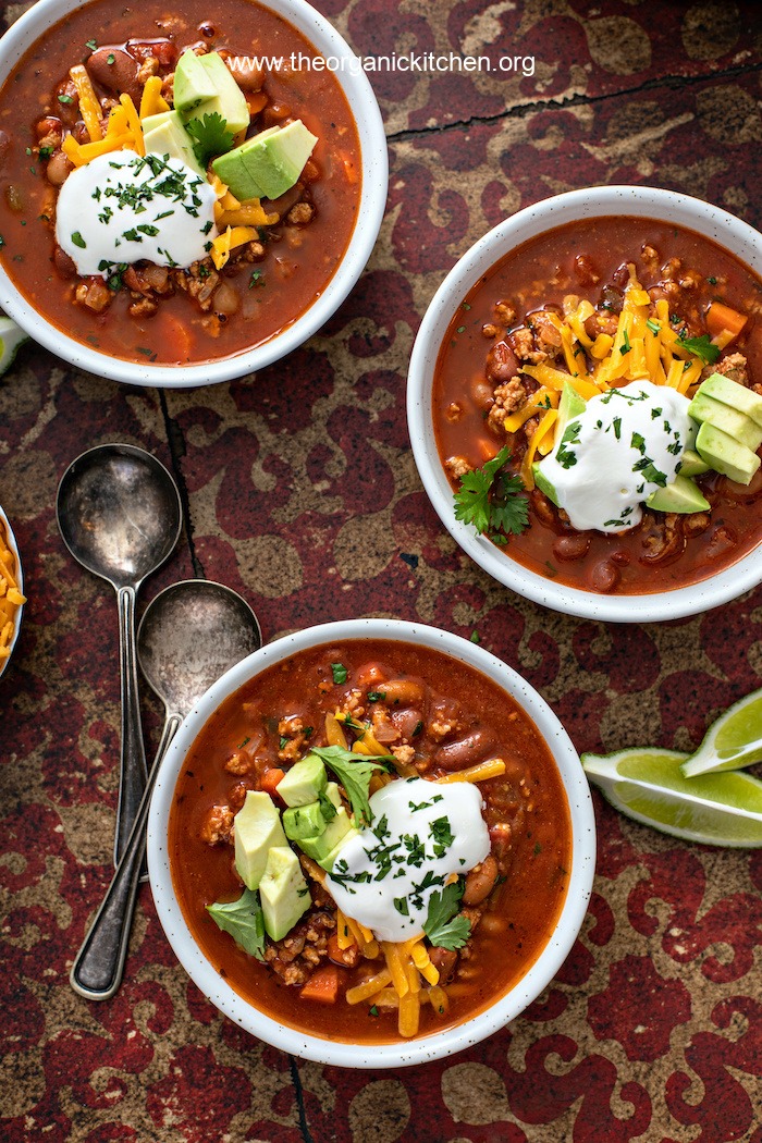 Flat lay of three bowls of 30 Minute Chipotle Turkey Chili garnished with cheddar cheese, sour cream and avocado , surrounded by lime wedges and silver utensils on a red and gold backdrop 