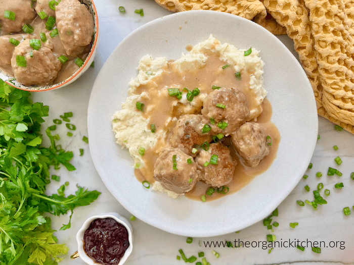 Swedish Meatballs with Lingonberry Jam and Mashed Potatoes on white plate set on white marble table, surrounded by herbs, a small bowl of jam and a bowl of meatballs