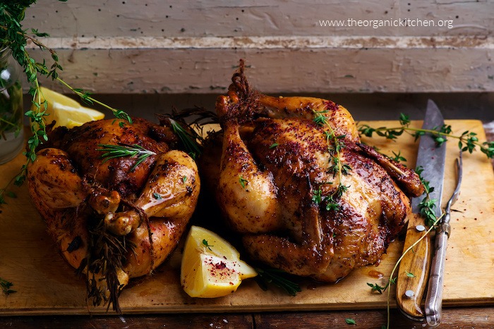 Two roast chickens on wood carving board with lemon wedges and carving utencils