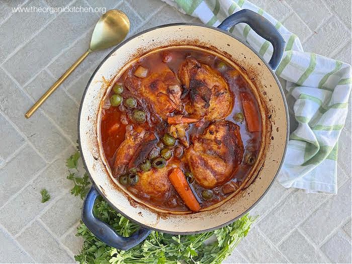 Dutch Oven Spanish Style Chicken on brick surface, surrounded by parsley, a dish towel and gold serving spoon