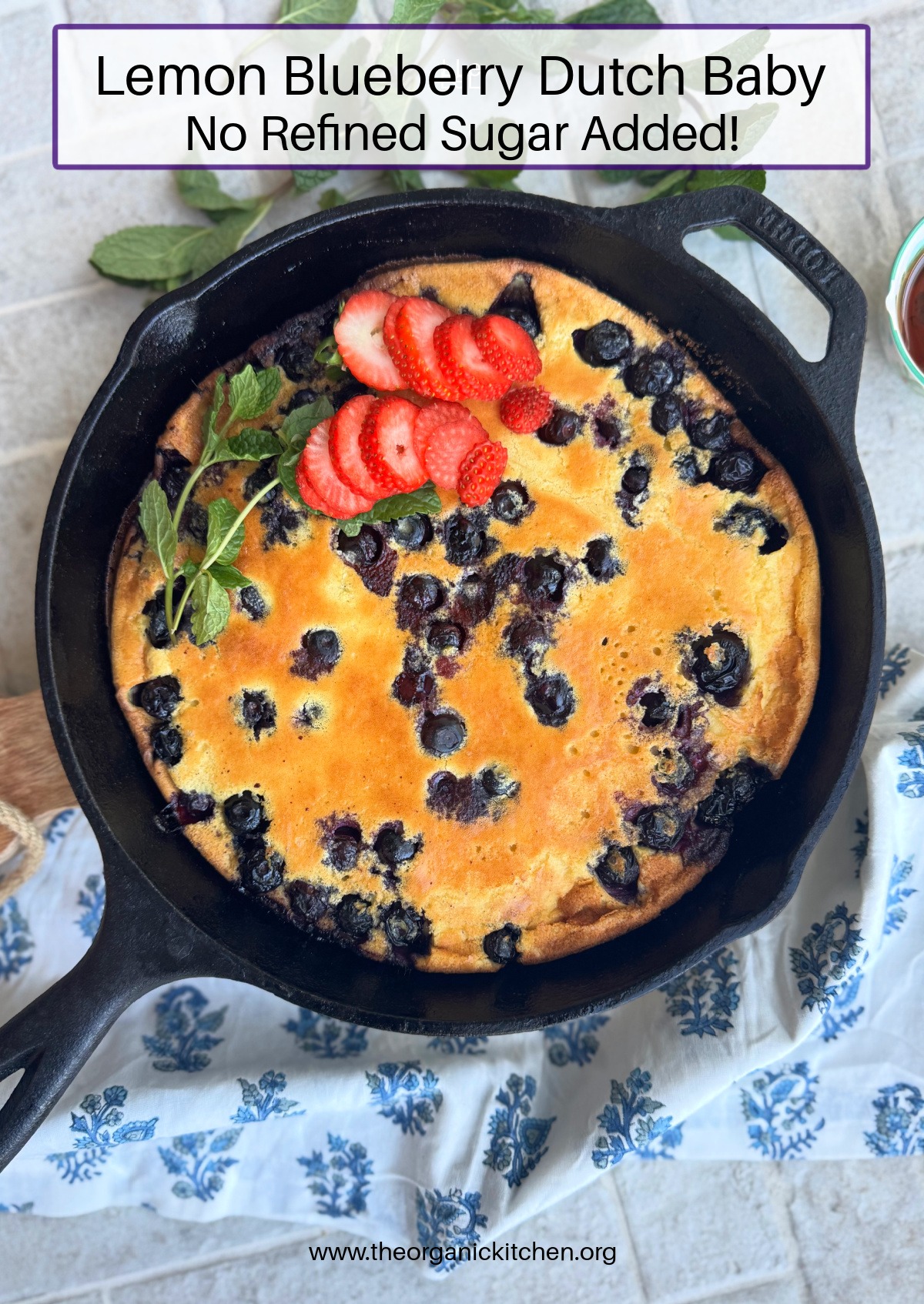Lemon Blueberry Dutch Baby (No Refined Sugar!) garnished with strawberries and mint in a black cast iron skillet set on gray bricks