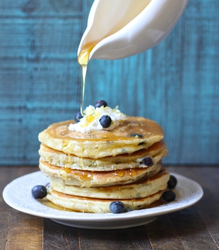 A stack of Blueberry Ricotta Pancakes on white plate with syrup being poured over from a white dish