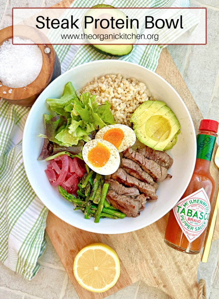 The Steak Protein Bowl in white bowl set on wood plank, surrrounded by a bottle of hot sauce, a lemon wedge, an avocado half, and a bowl of sea salt