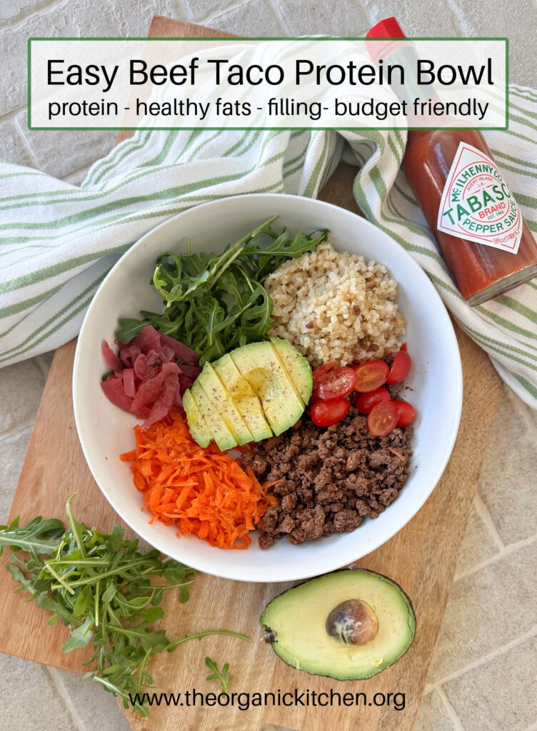 A white bowl on a wooden platter filled with ground beef, veggies, rice and avocado surrounded by a green and white dish twoel