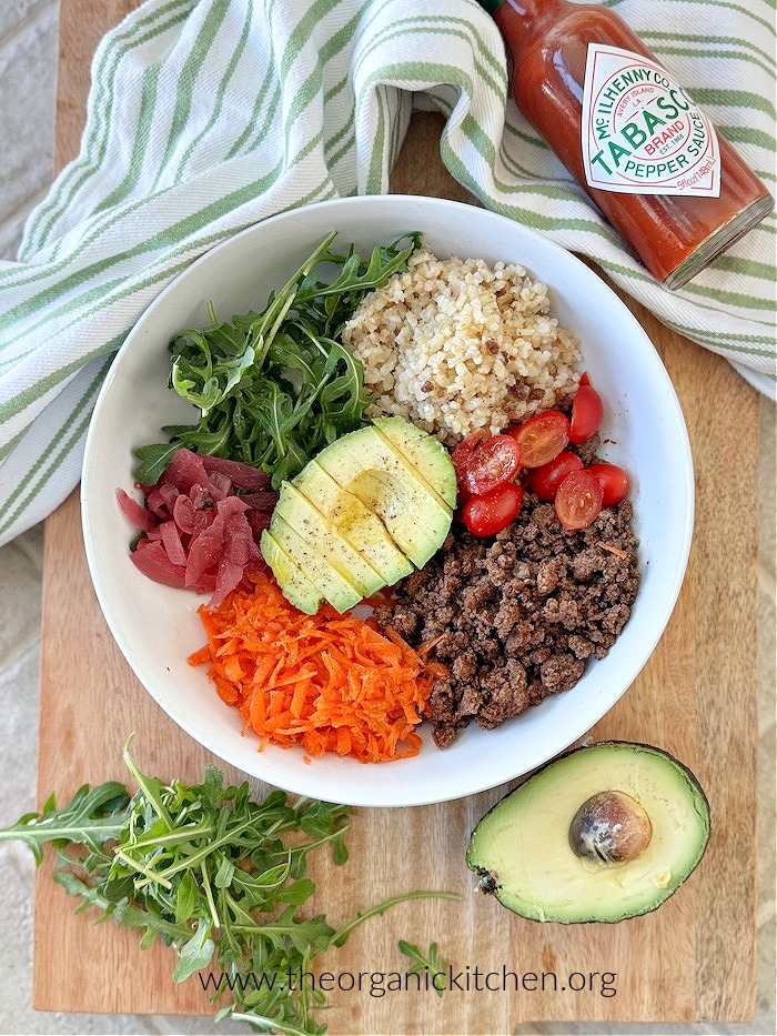Easy Beef Taco Protein Bowl in white bowl on wood board with half an avocado, tabasco and green and white dish towel