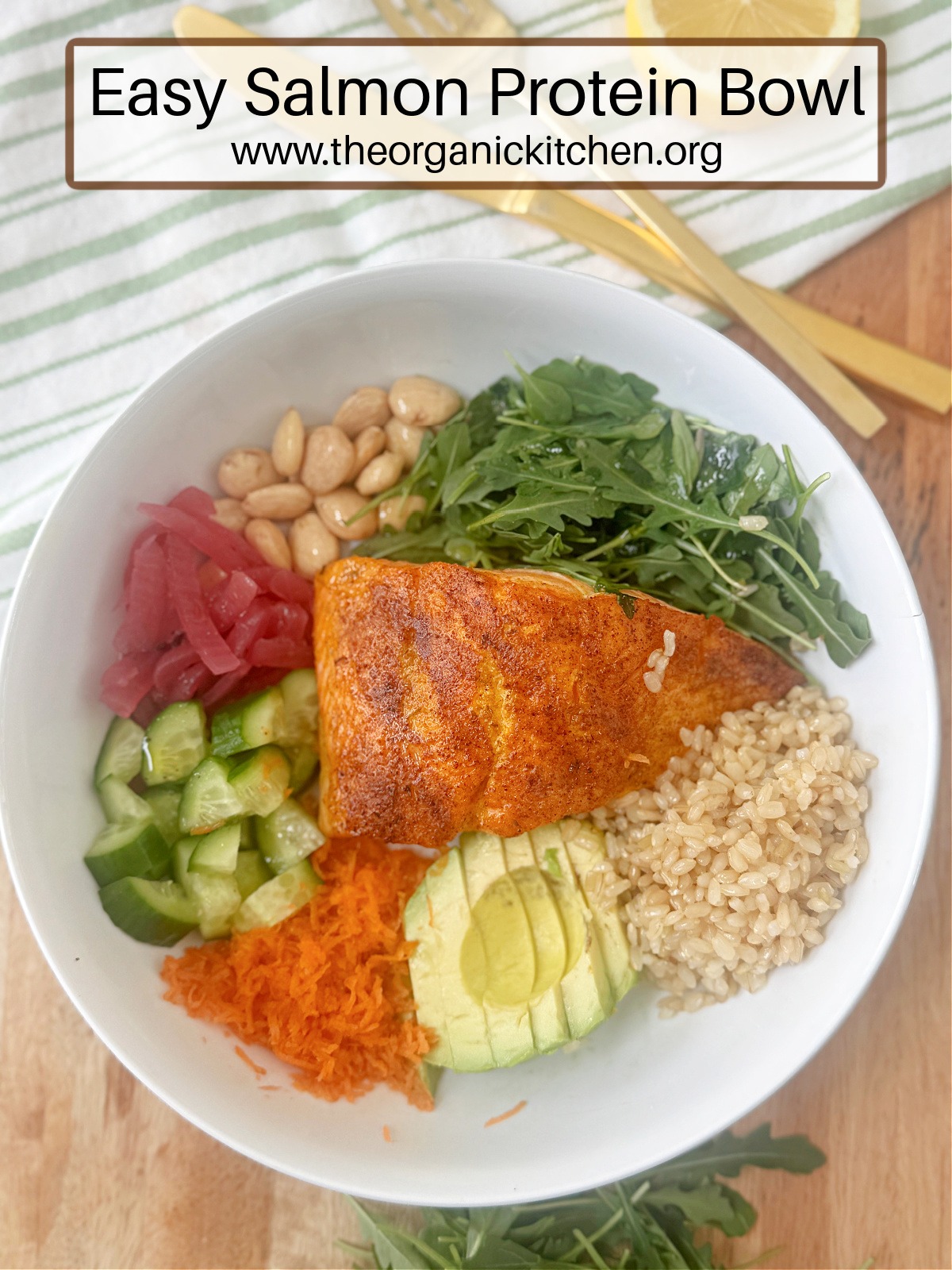 Easy Salmon Protein Bowl: a white bowl with salmon, arugula, red onions, cucumbers, brown rice, and avocado on wood platter