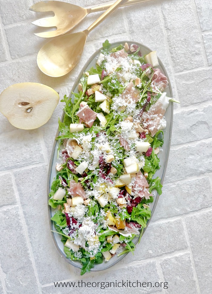 The Italian Salad on gray platter set on brick background with gold tongs, half a pear and dish towel in background