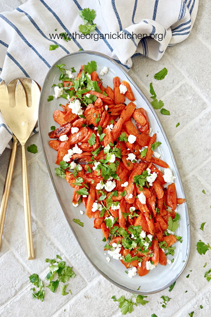 Roasted Maple Glazed Carrots with Feta on gray platter, set on gray bricks with gold serving utensils and dish towel in background