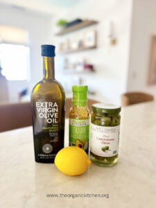 A bottle of olive oil, Italian dressing, a jar of olives, and a fresh lemon sitting on a countertop