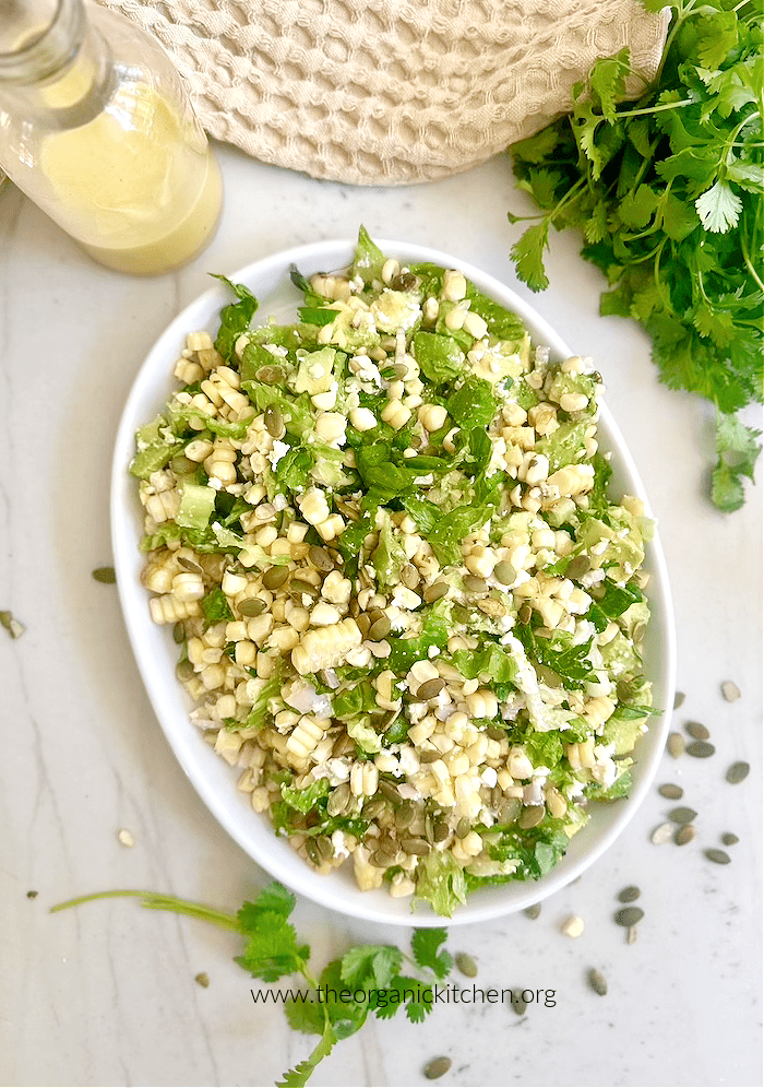 Grilled Corn and Avocado Salad on white platter, surrounded by herbs and pumpkin seeds, with a bottle of salad dressing