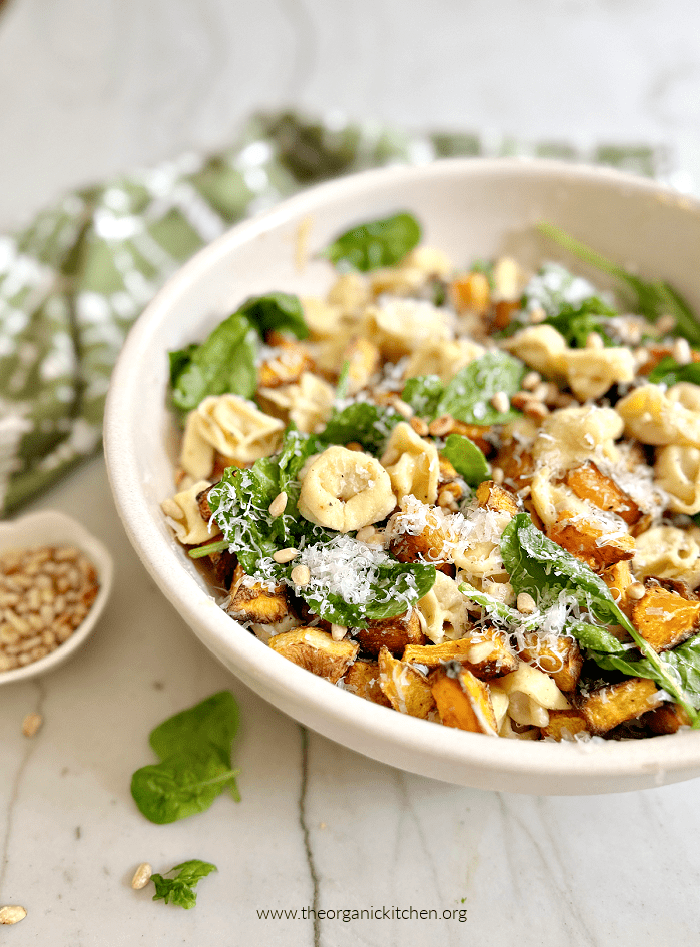 A white bowl filled with Browned Butter Tortellini with Roasted Butternut Squash, with a dish towel , a bowl of pine nuts, and spinach leaves scattered around