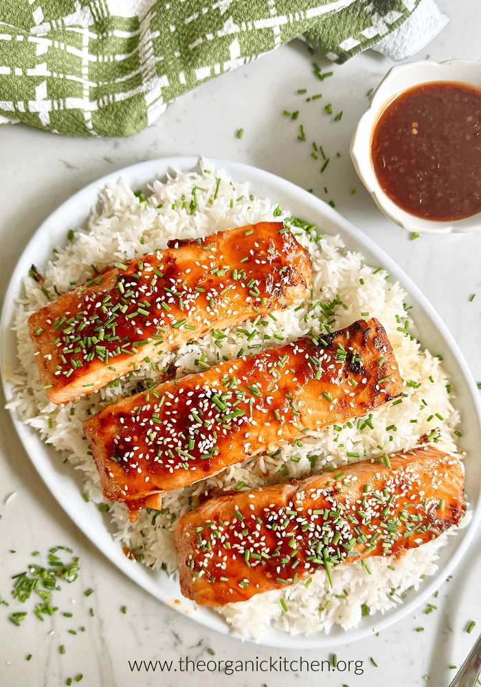 Apricot Teriyaki Salmon on white rice served on white plate, with green dish towel and small bowl of sauce on the side