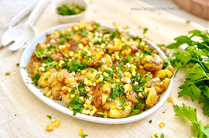 Crispy Smashed Potato and Corn Salad grnished with herbs on white platter with a bowl of herbs and serving utensils in the background