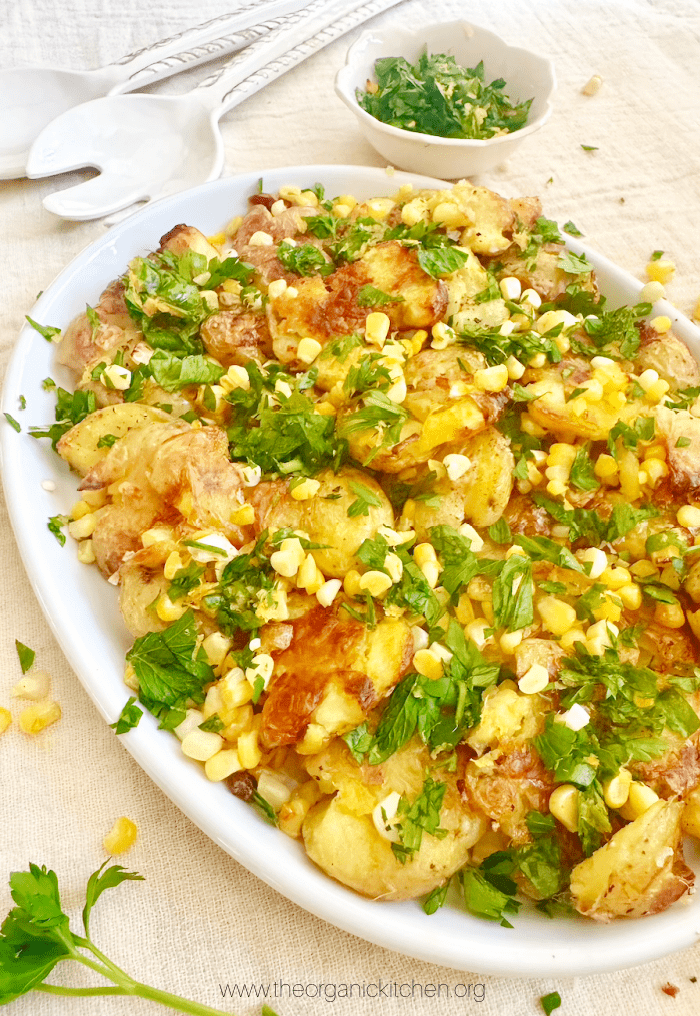 Crispy Smashed Potato and Corn Salad garnished with gremolata on white serving dish with a bowl and herbs in the background