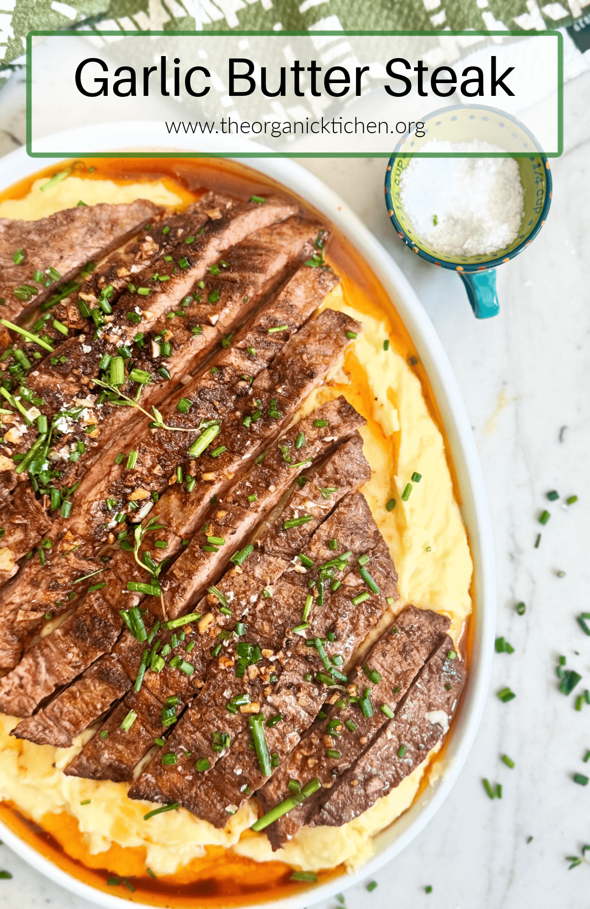 Garlic Butter Steak atop mashed potatoes on white plate with a small bowl of sea salt and green and white dish towel above