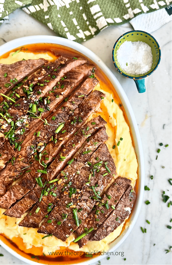 Garlic Butter Steak on mashed potatoes, garnished with chives with a small green bowl of sea salt and dishtowel above