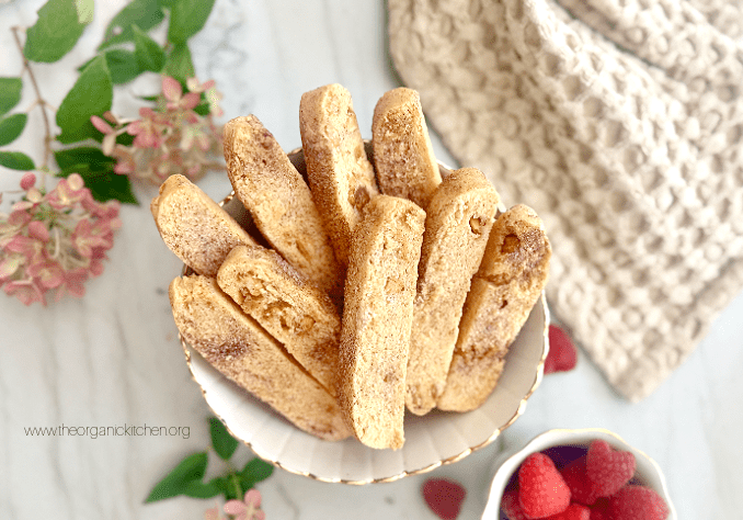 White Chocolate Biscotti stacked in a bowl with hydrangea, raspberries, and a beige dish towel in background