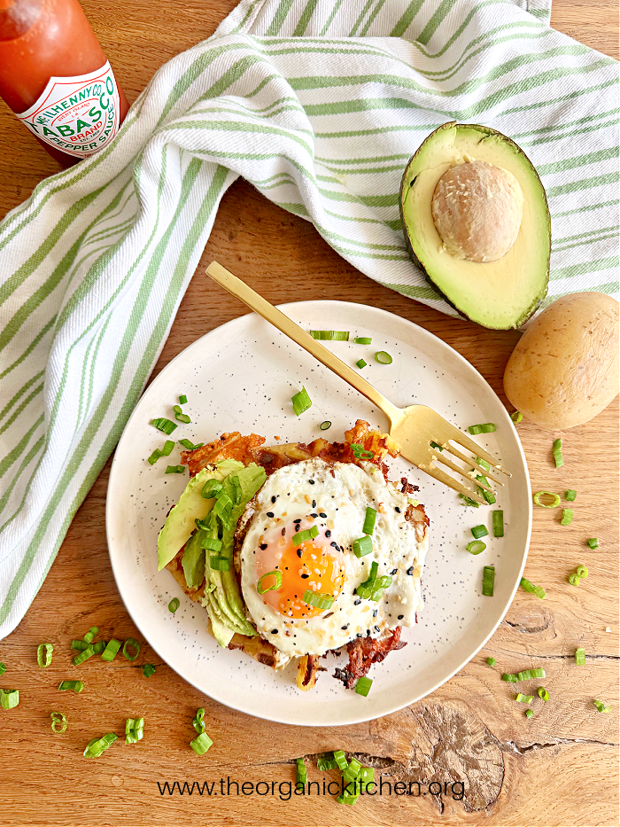 Parmesan Potato Waffle with fried egg, avocado, and green onions on white plate with gold fork and green and white dish towel